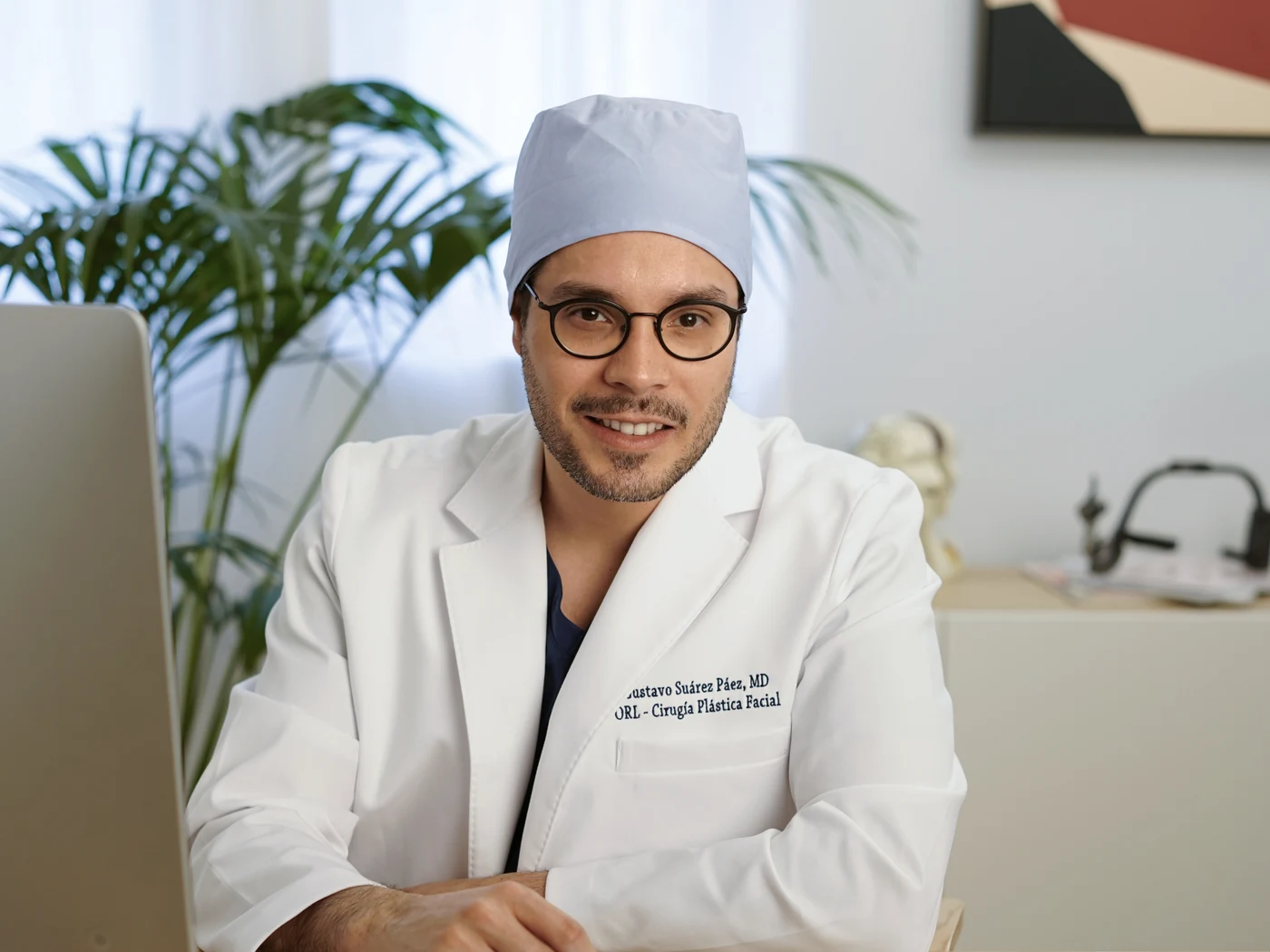 Dr. Gustavo Suárez, in a scrub cap and white coat, smiling at the camera at his consulting desk