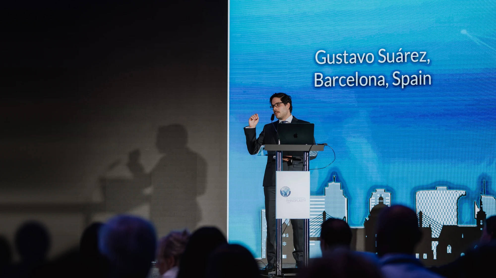 El Dr. Gustavo Suárez durante una ponencia en un congreso, junto al atril, con su nombre proyectado en la pantalla azul